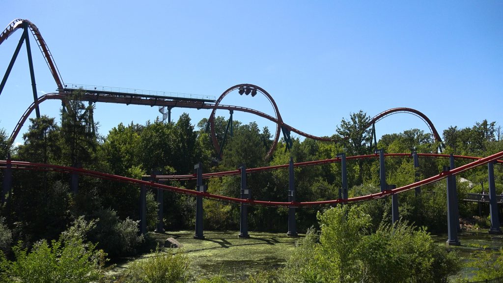 A train at the top of Yukon Striker's vertical loop. It's foregrounded by many trees and the lake under Vortex (Lake LeBarge).